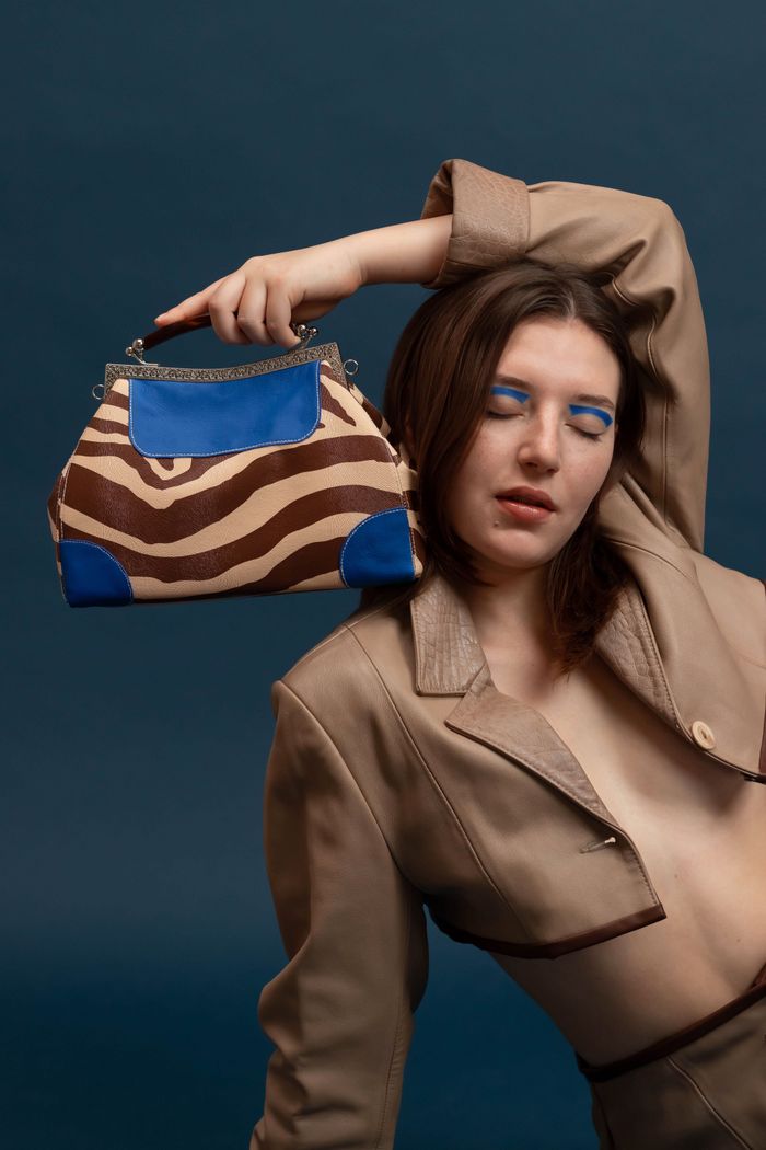 about-us-01 A well-dressed woman poses with a stylish handbag against a blue background, showcasing fashion.
