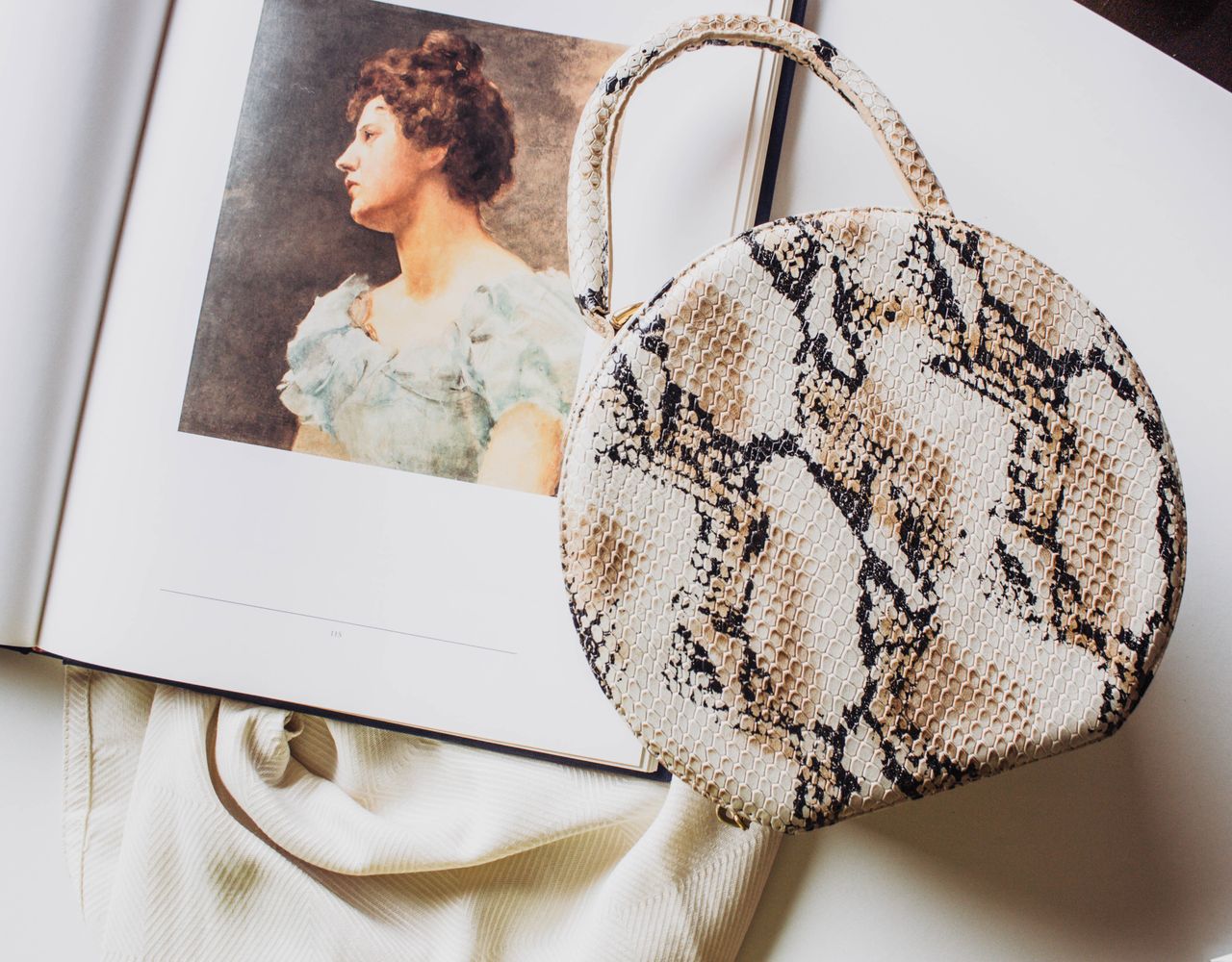 heros-img Round snakeskin handbag placed on an open art book with a portrait.